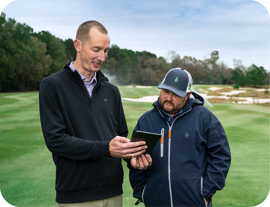 A man with short hair in a dark blue pullover half-zip sweater, assisting his colleague in a dark blue hat and zip-up rain jacket. The man with short hair on the left is showing his colleague something on an iPad as he speaks.
