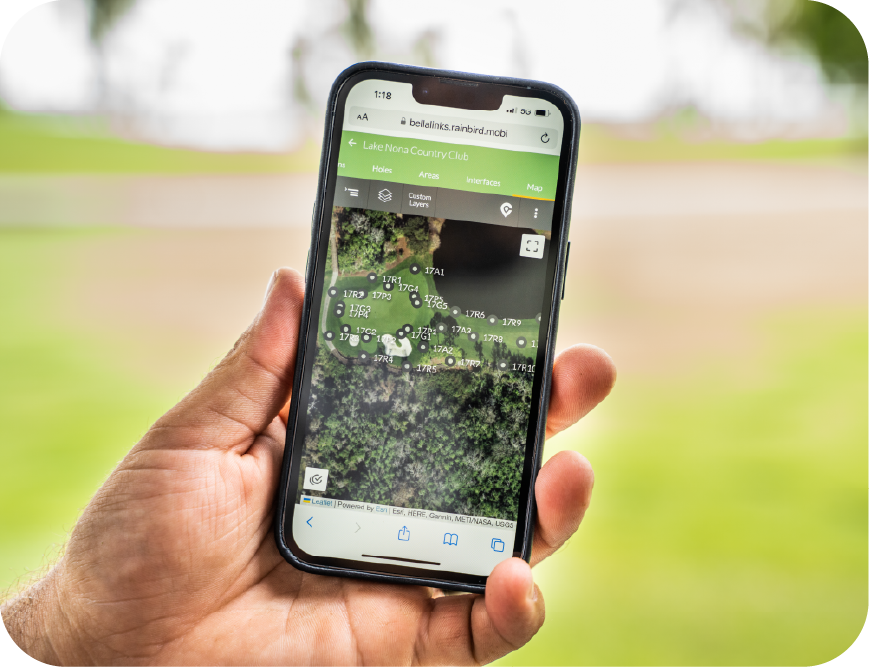 A hand holding a smartphone, showing a golf course irrigation map in overhead satellite view.