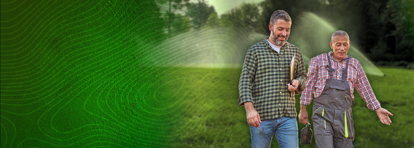 Landscape contractor walking alongside another man in a field with irrigation sprinklers shooting water in the background