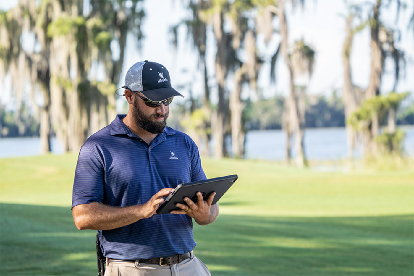 A man with a dark brown beard wearing a baseball cap, polo, and khakis, is holding a large iPad and making selections on the screen.